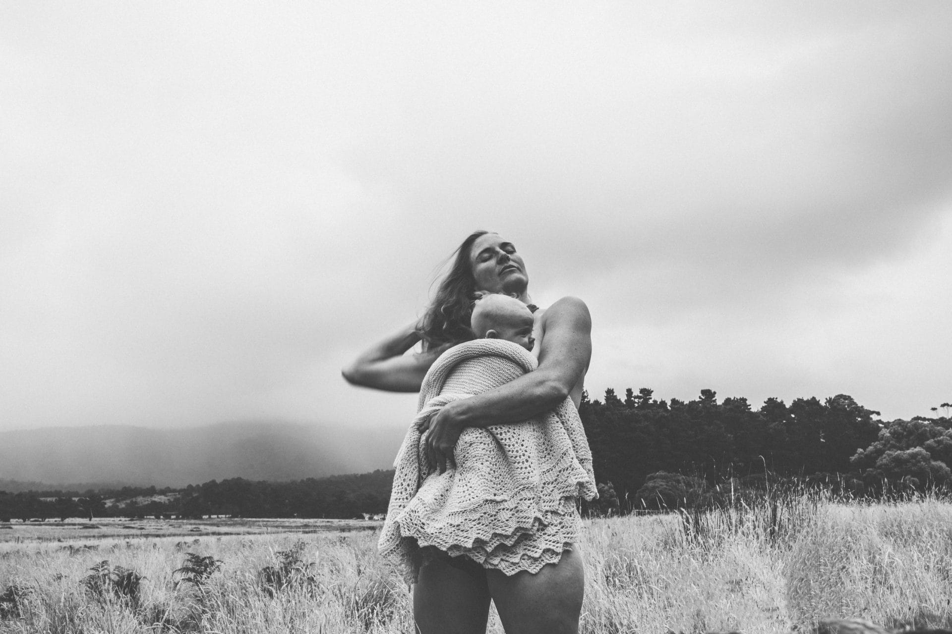 A woman in a knit dress stands in a field, embracing herself and looking upwards, with a forest and mountains in the background, all in black and white.