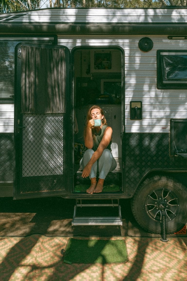 Natasha Grech sipping coffee while sitting in the doorway of a camper van parked in a wooded area, sunlight filtering through the trees.