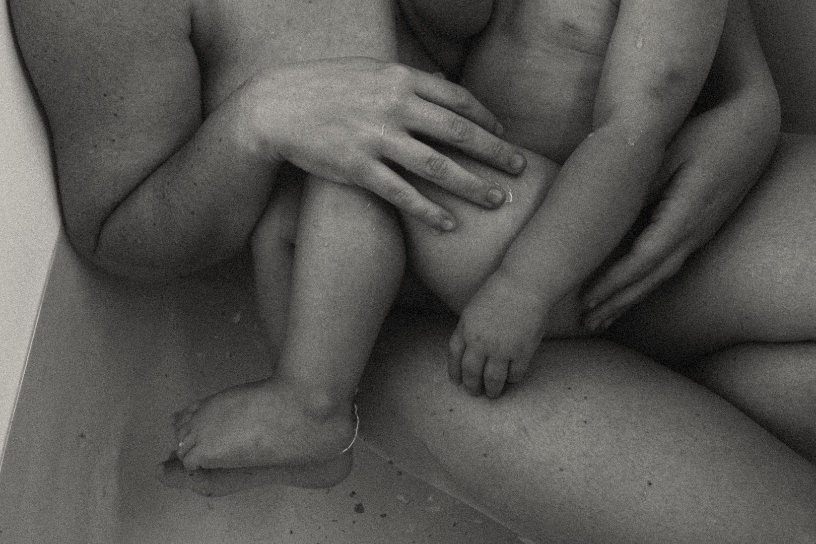 Black and white close-up of an adult and a child sitting in a bath, focusing on their overlapping arms and legs.