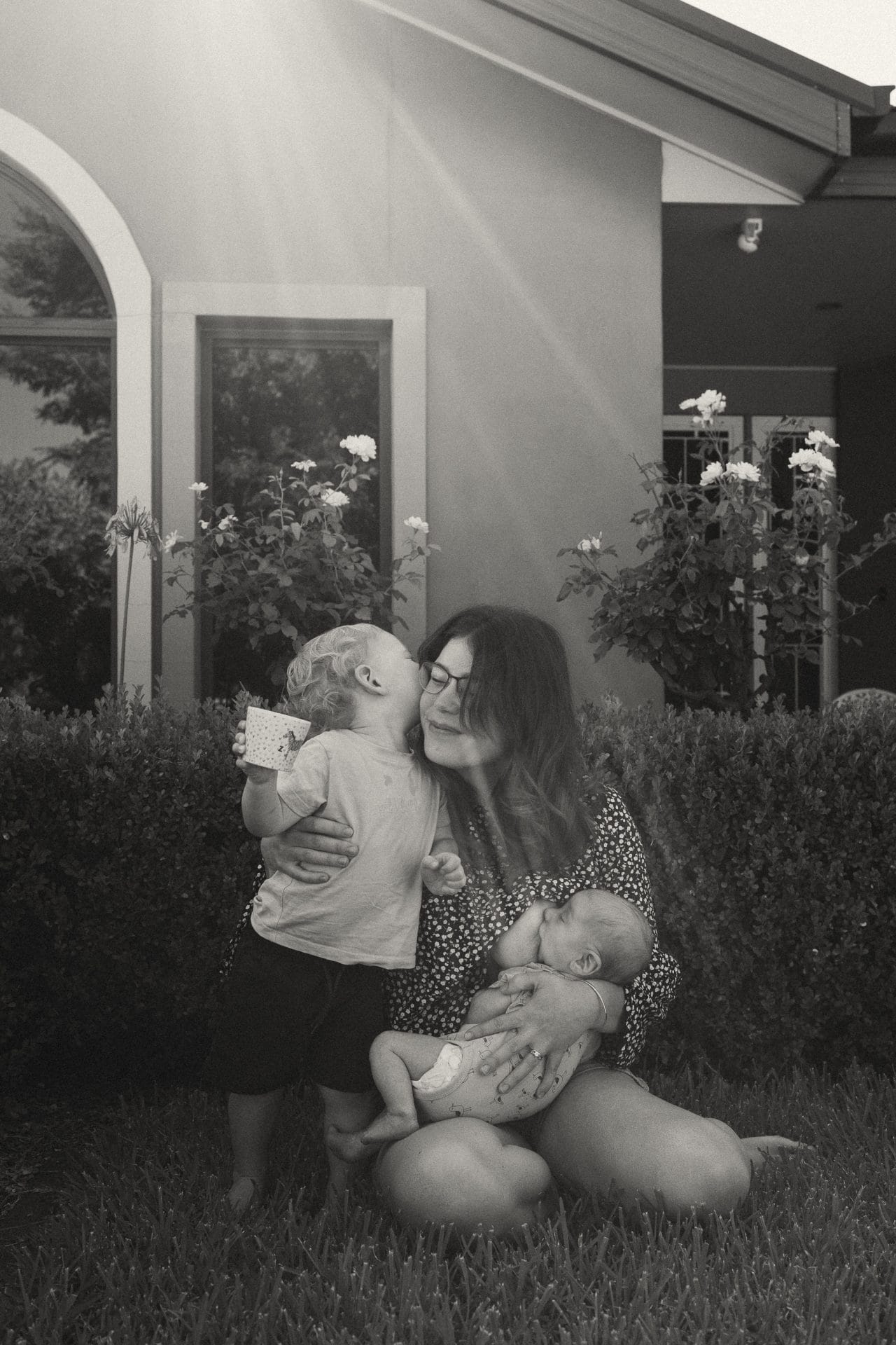 A woman kneeling smiles while holding a baby and looking at a toddler holding a cup in a garden, with sunlight filtering through.