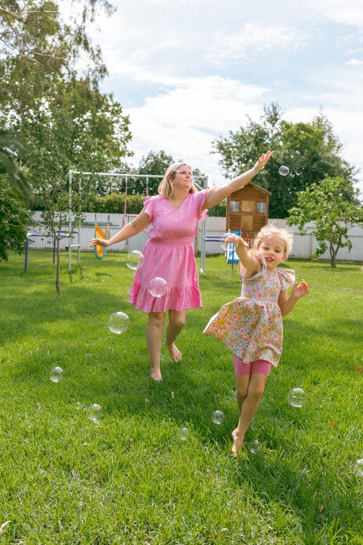 A woman and a young girl joyfully running through a grassy yard with soap bubbles, with playground equipment in the background.
