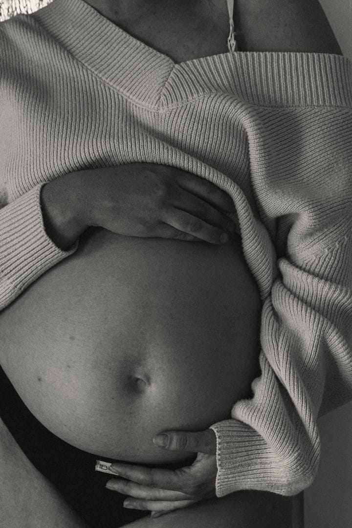 Black and white image of a pregnant woman cradling her belly, wearing an off-shoulder sweater. hands visible, intimate and gentle touch.