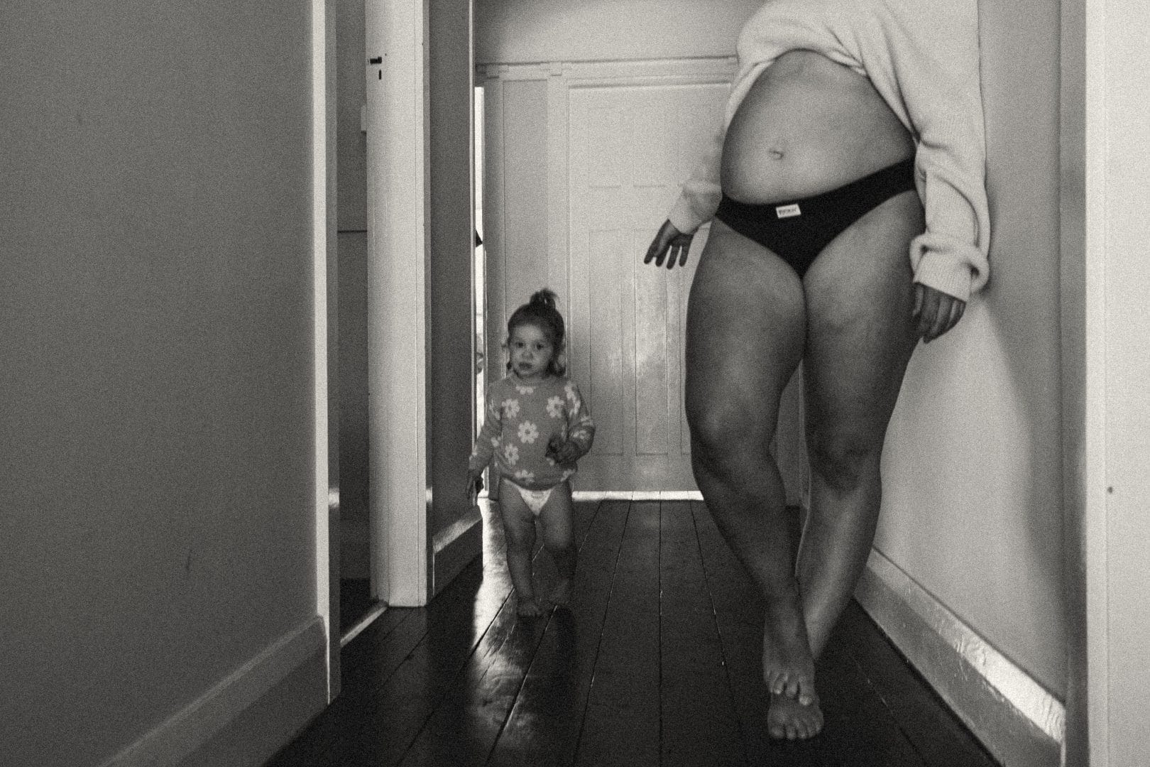 Black and white photo of a pregnant woman and a toddler walking down a hallway.