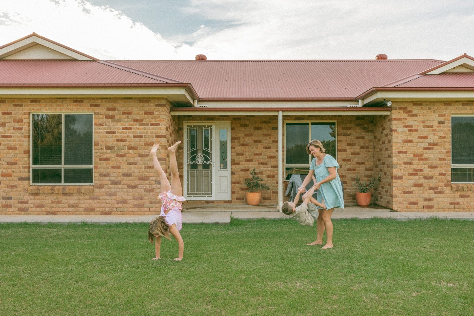 A woman and a young girl playing with a ball on the lawn in front of a brick house; the girl is doing a cartwheel.