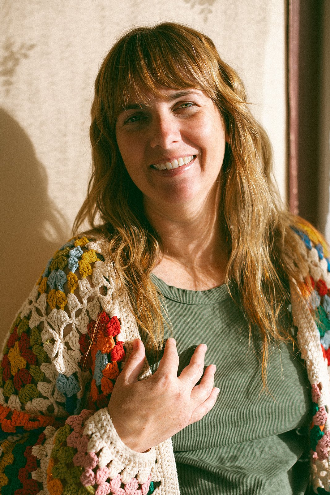 A person with long hair smiles while wearing a colorful knitted sweater and a green shirt.