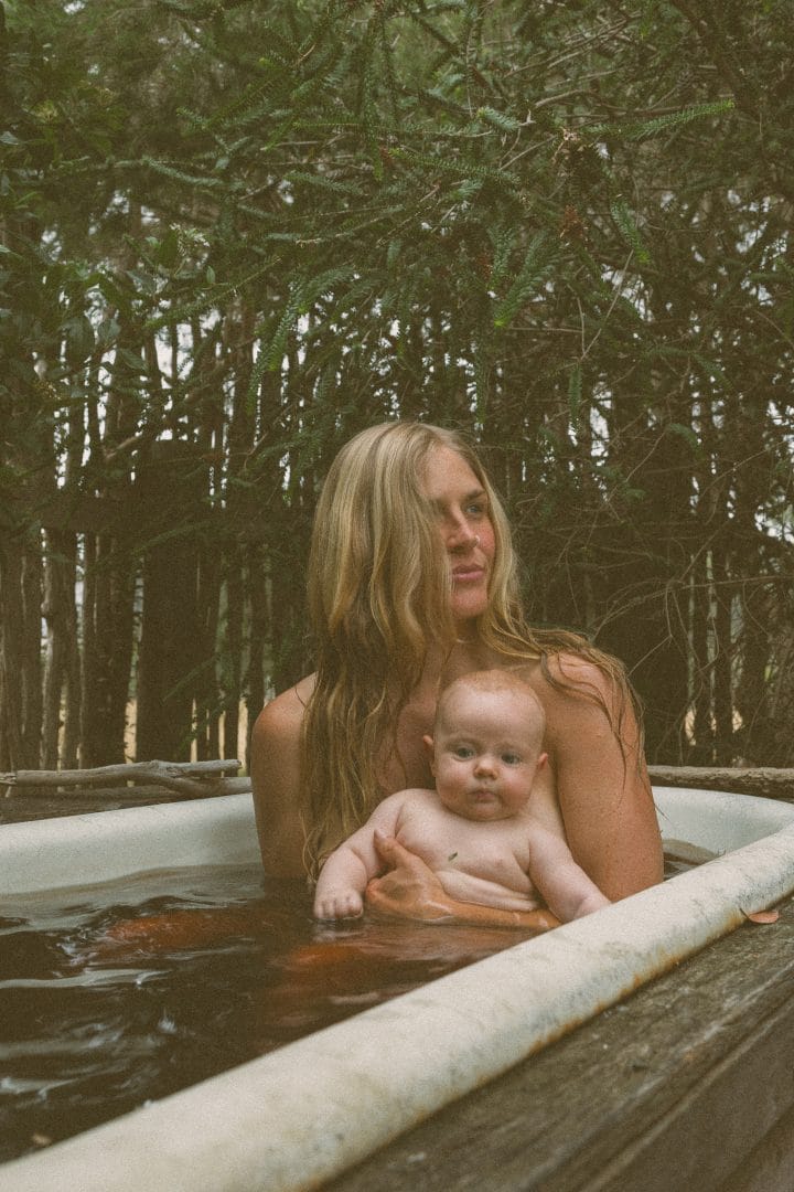 A woman holding a baby in an outdoor wooden tub surrounded by trees.