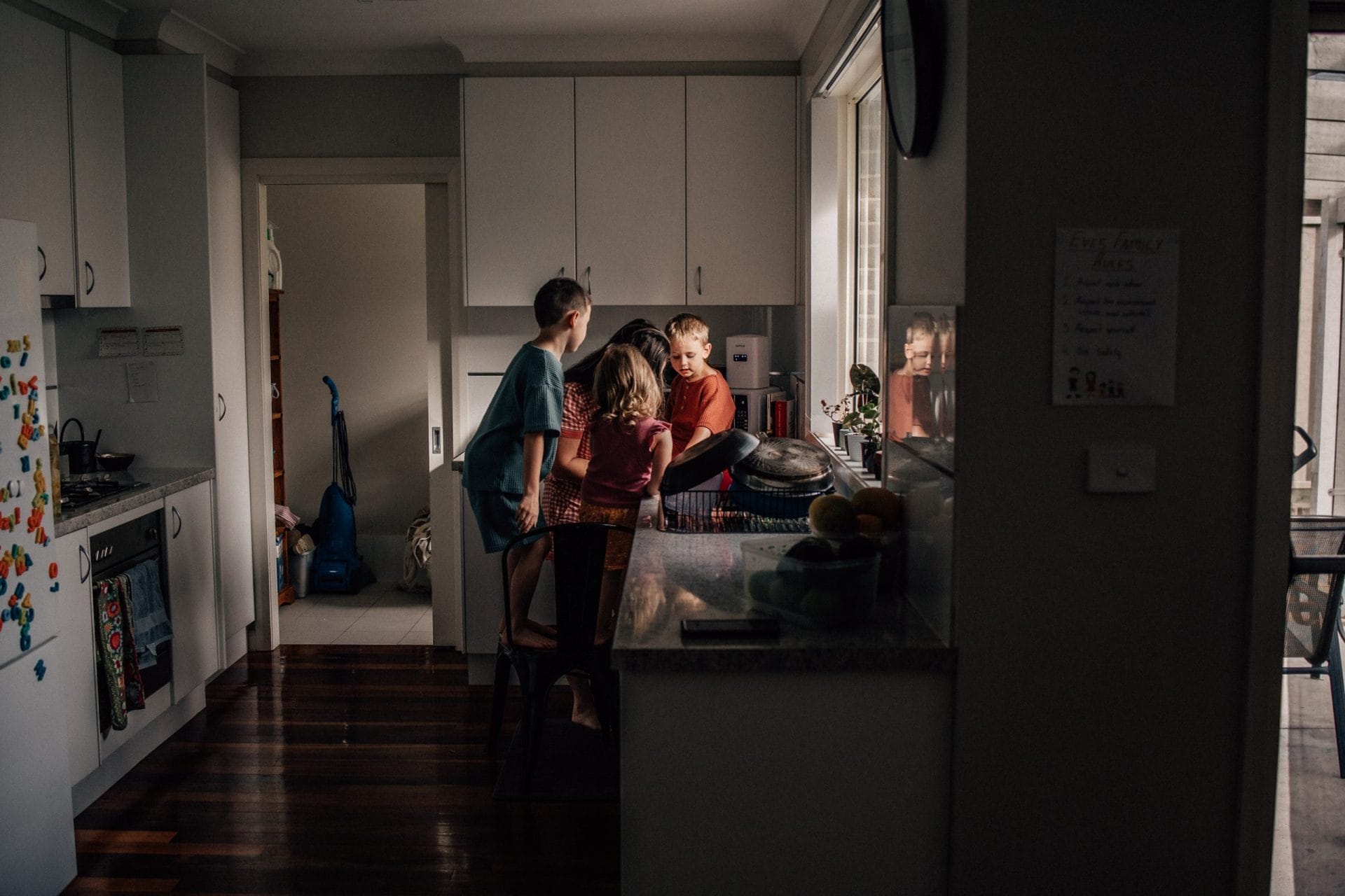Two children and an adult in a kitchen, engaging in conversation by a counter with fruit bowl, in soft natural light.