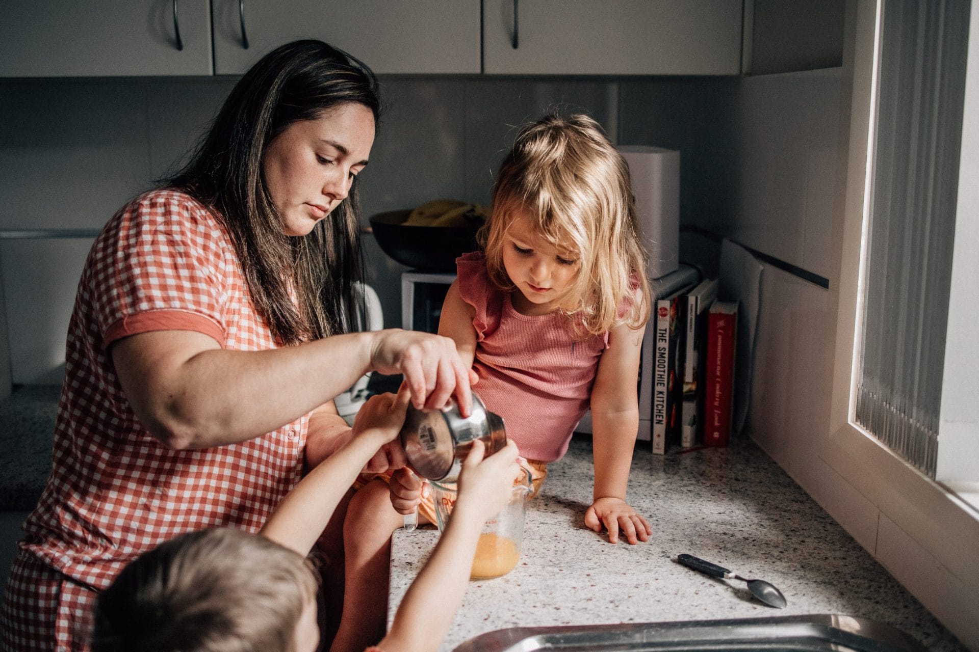 The need for perfection is stealing our joy 3 A woman and two children baking in a kitchen, with the woman helping a young girl pour ingredients into a bowl.