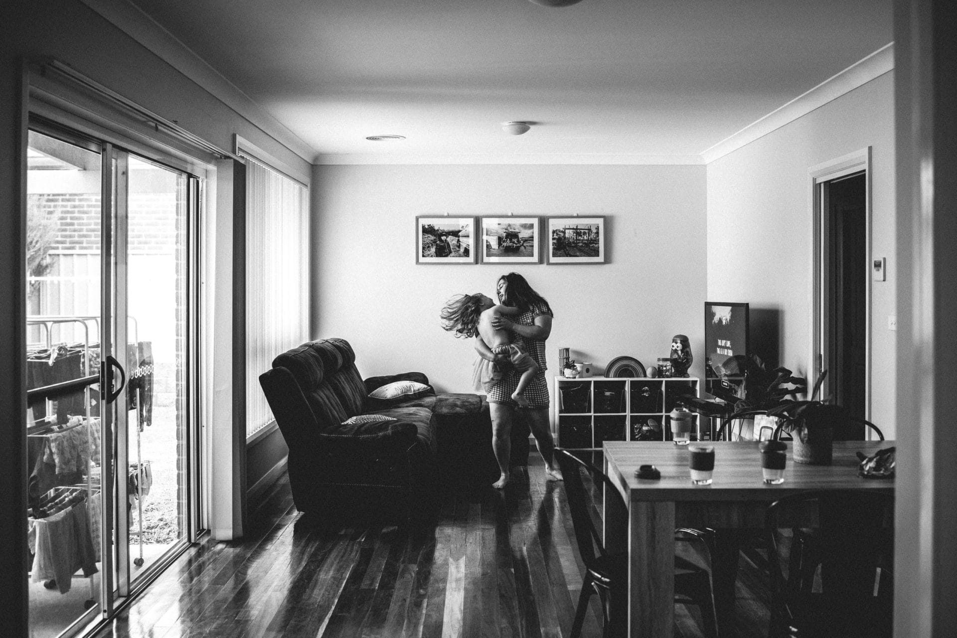 A black and white photo of a woman holding a child in her arms, spinning around joyfully in a living room with sliding doors open to a backyard.