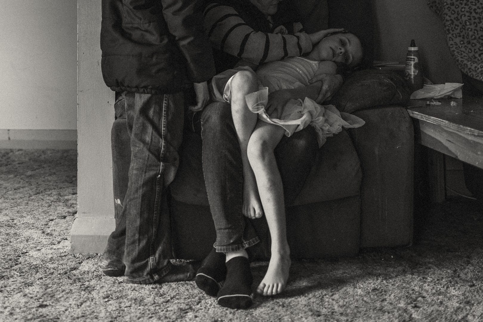 A black and white photo of a child sleeping on a lap with two men standing nearby, one touching her foot, in a modestly furnished room.