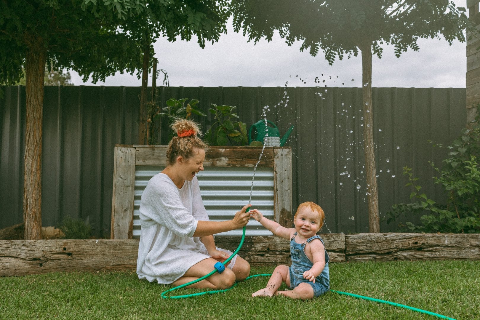 A woman and a toddler play with a garden hose on the grass in a backyard, smiling and enjoying the spraying water.