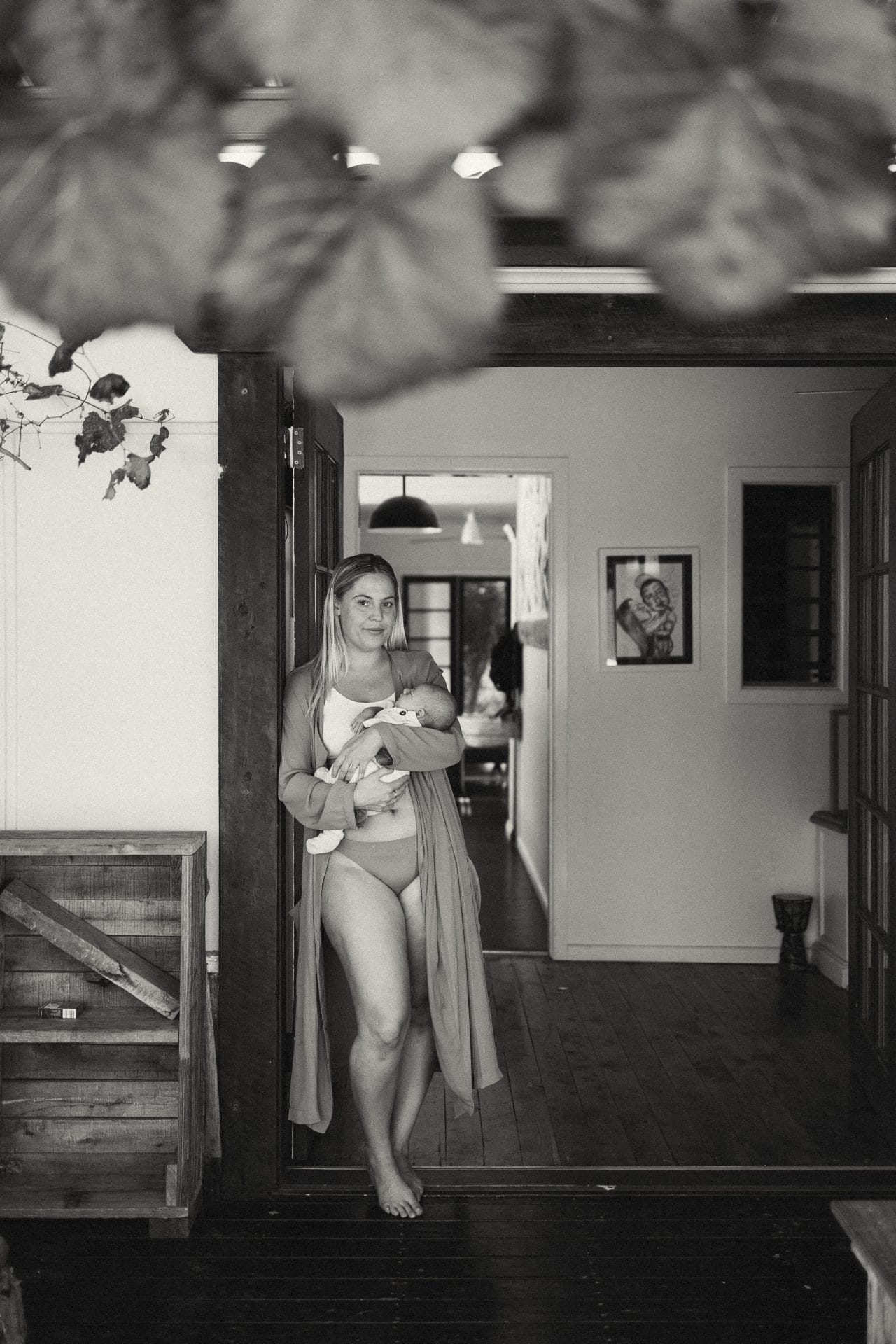 A mother, wrapped in a light shawl, stands barefoot in a doorway cradling her baby, with a softly lit interior in the background.