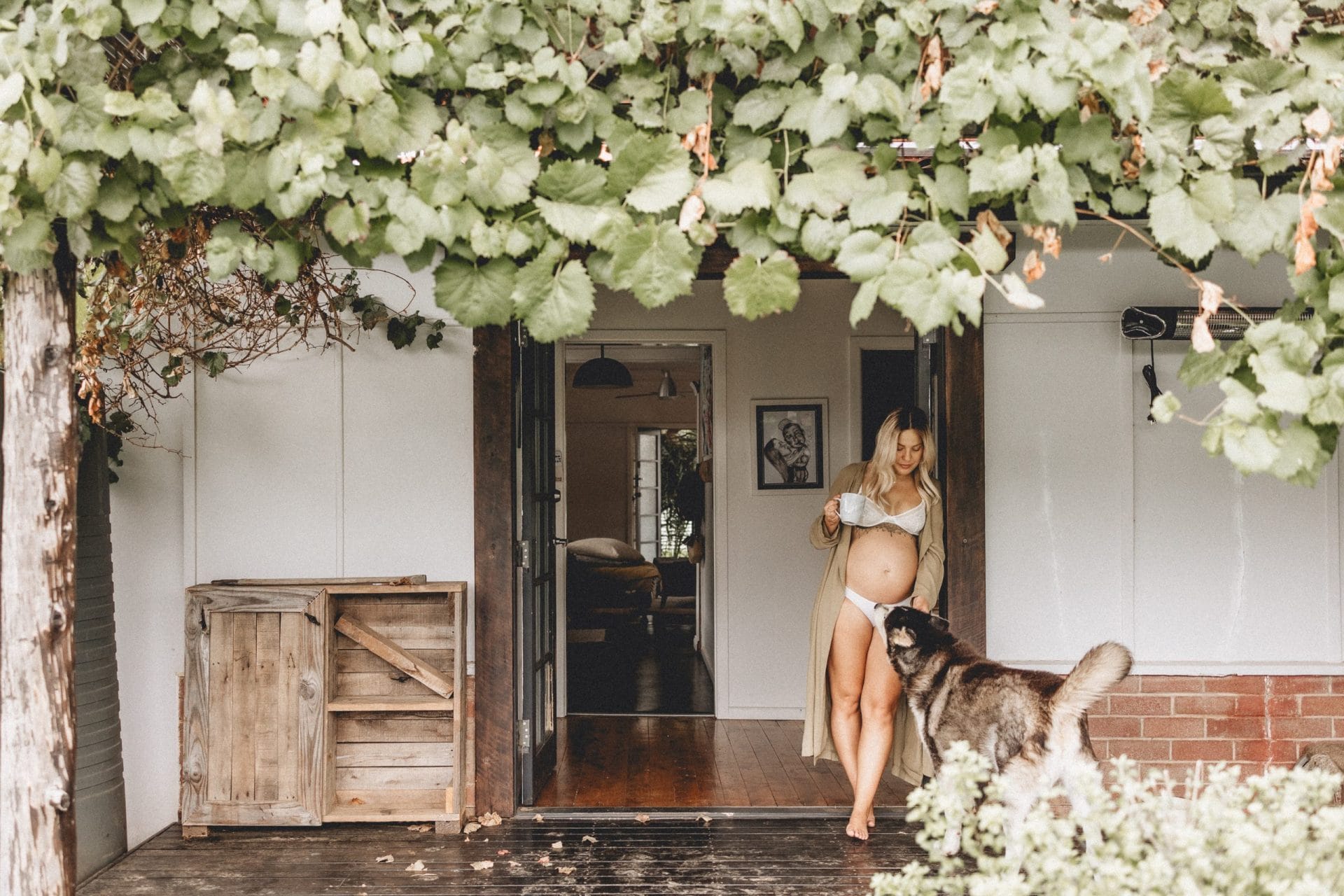 Pregnant woman standing at her house entrance under a vine canopy, stroking a dog, in a serene, rustic setting.
