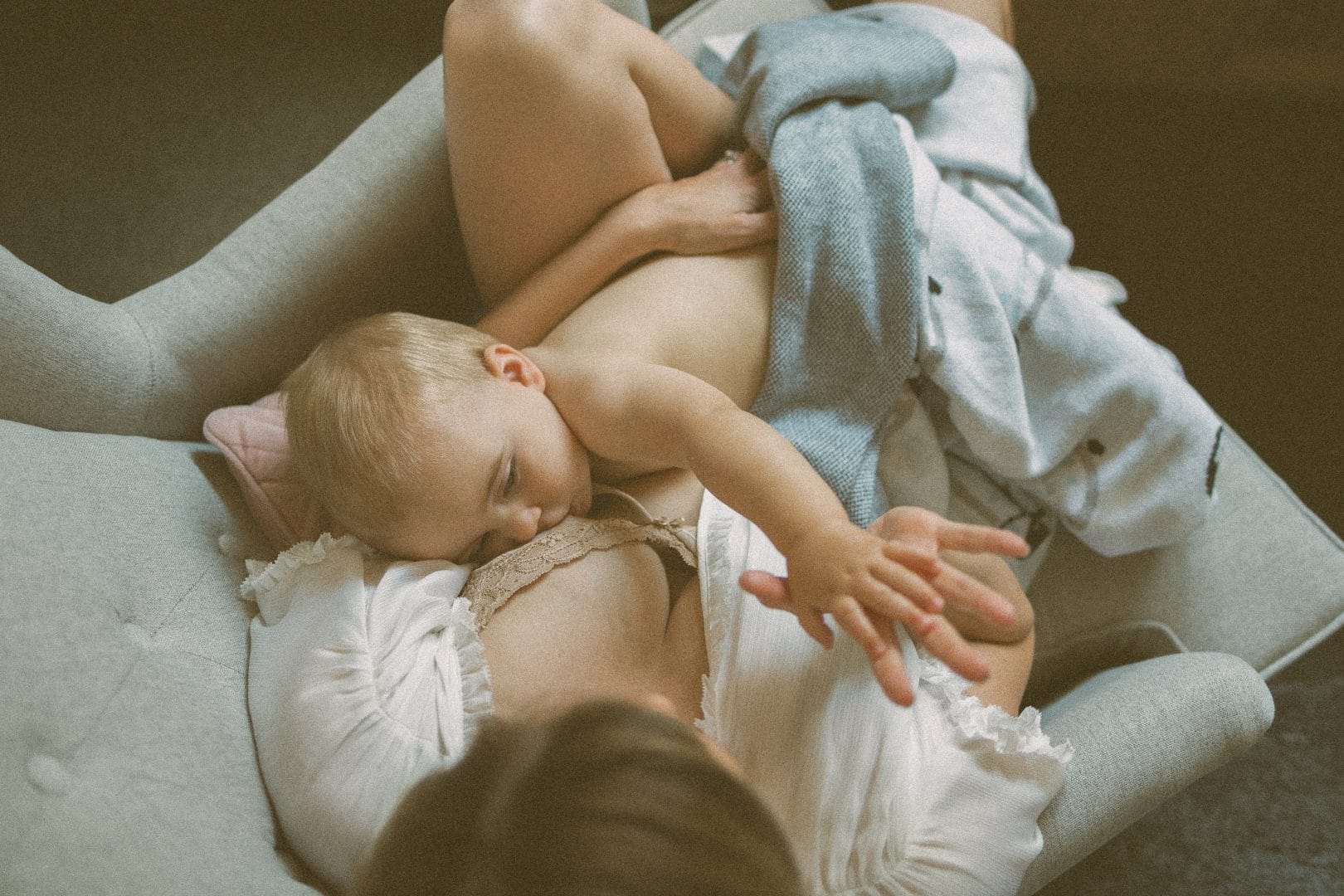 A mother in a white blouse cradling a sleeping baby on a grey sofa, covered partially by a blue blanket.