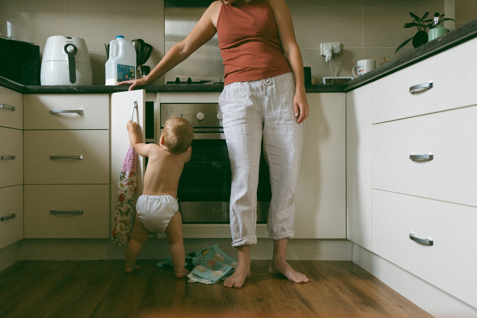 A toddler in a diaper reaches for a drawer in a kitchen, standing next to an adult woman in casual clothes who is supervising.