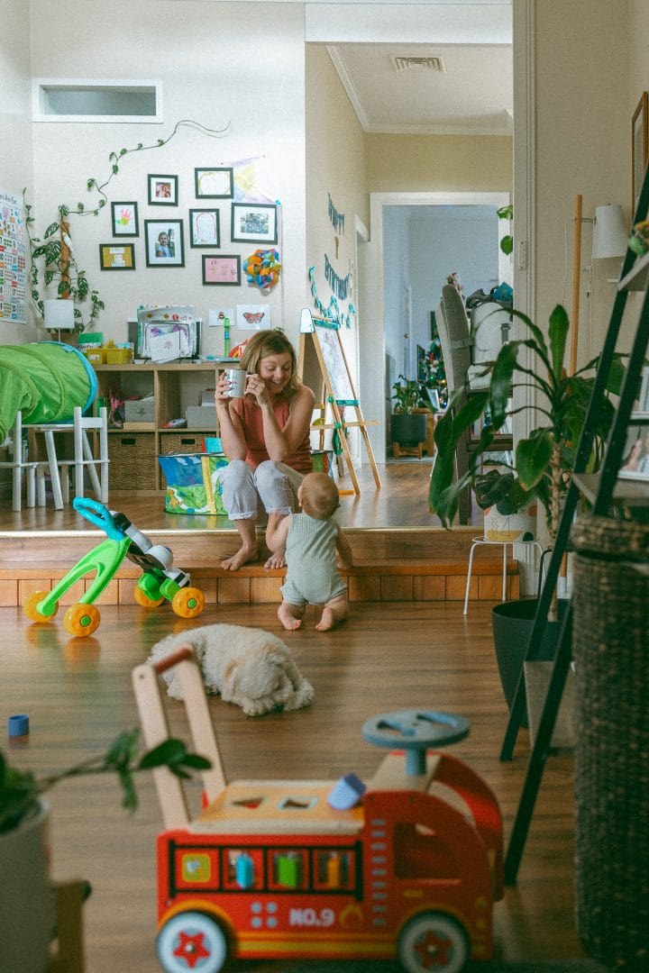 A woman using a smartphone sits on stairs in a colorful home interior, surrounded by children's toys and a dog, while a child stands beside her.