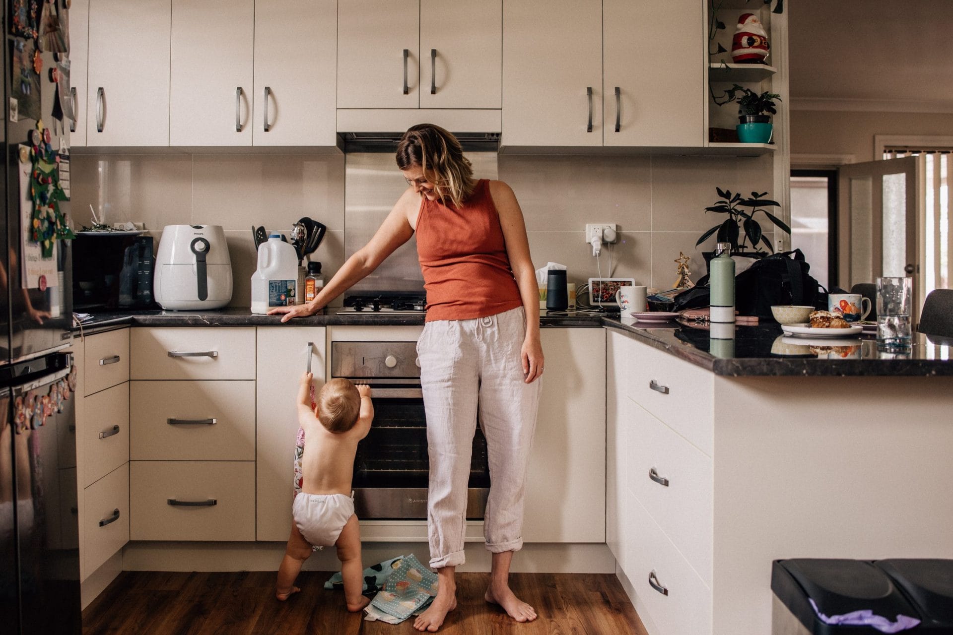 A mother and her toddler stand by an open oven in a cluttered kitchen, with the child reaching up towards the oven.