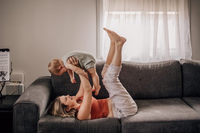 A woman lying on a couch lifts a young child with her feet, both smiling in a warmly lit living room.