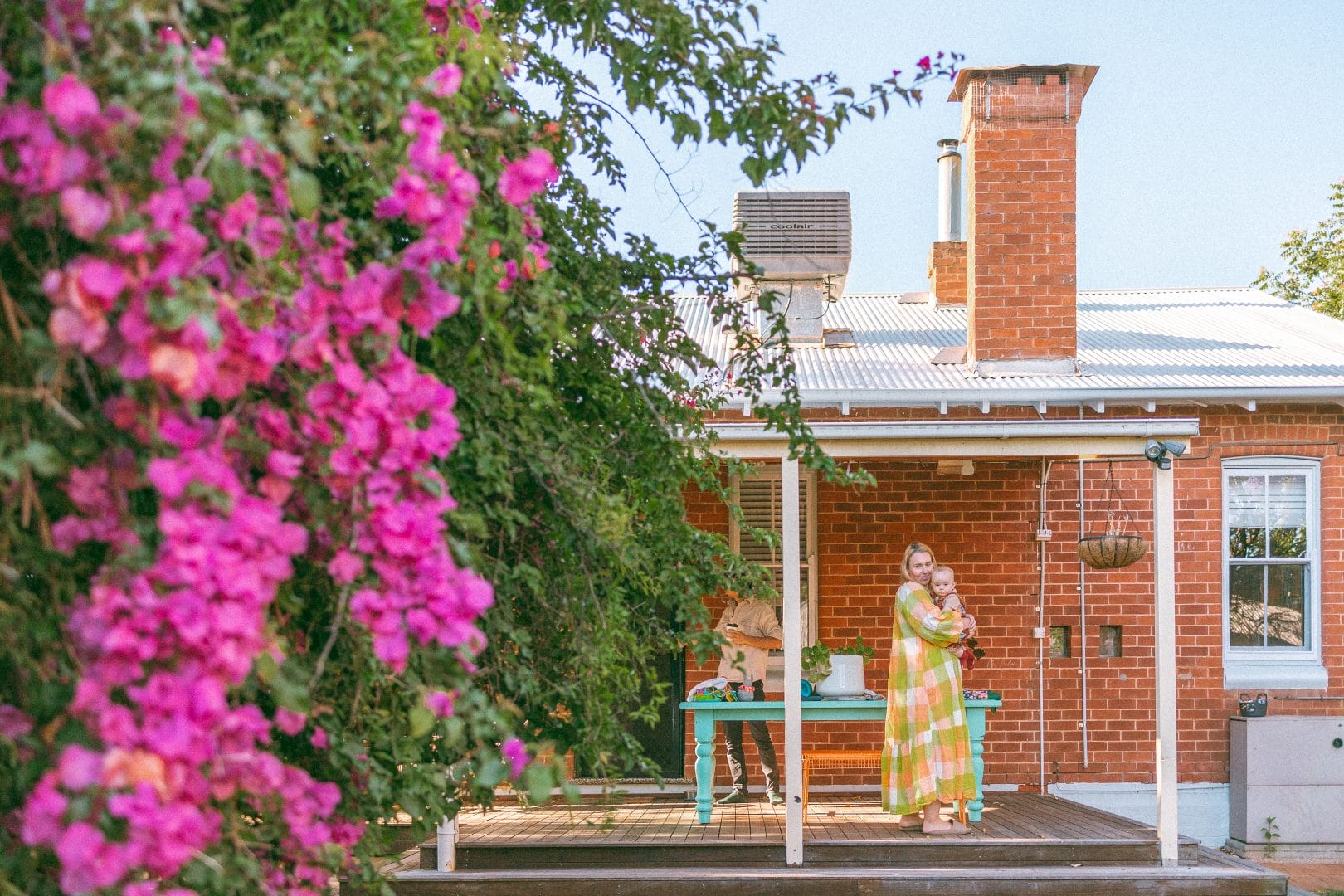 A woman and child on a sunlit porch of a brick house, surrounded by vibrant bougainvillea, engaging in an activity at a table.