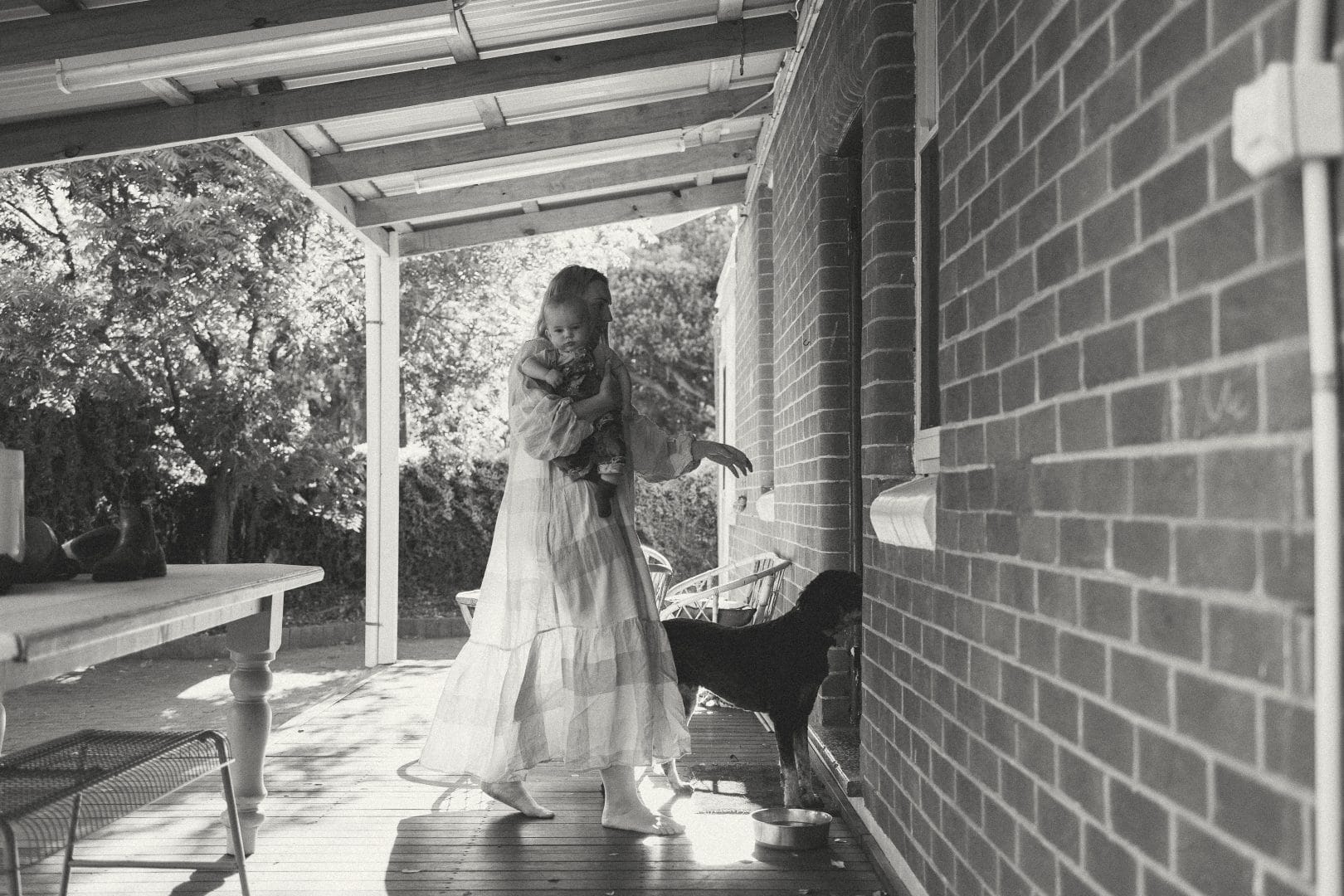 A young girl in a flowing dress holds a cat on a shaded porch next to a brick wall, with outdoor furniture visible in the background.