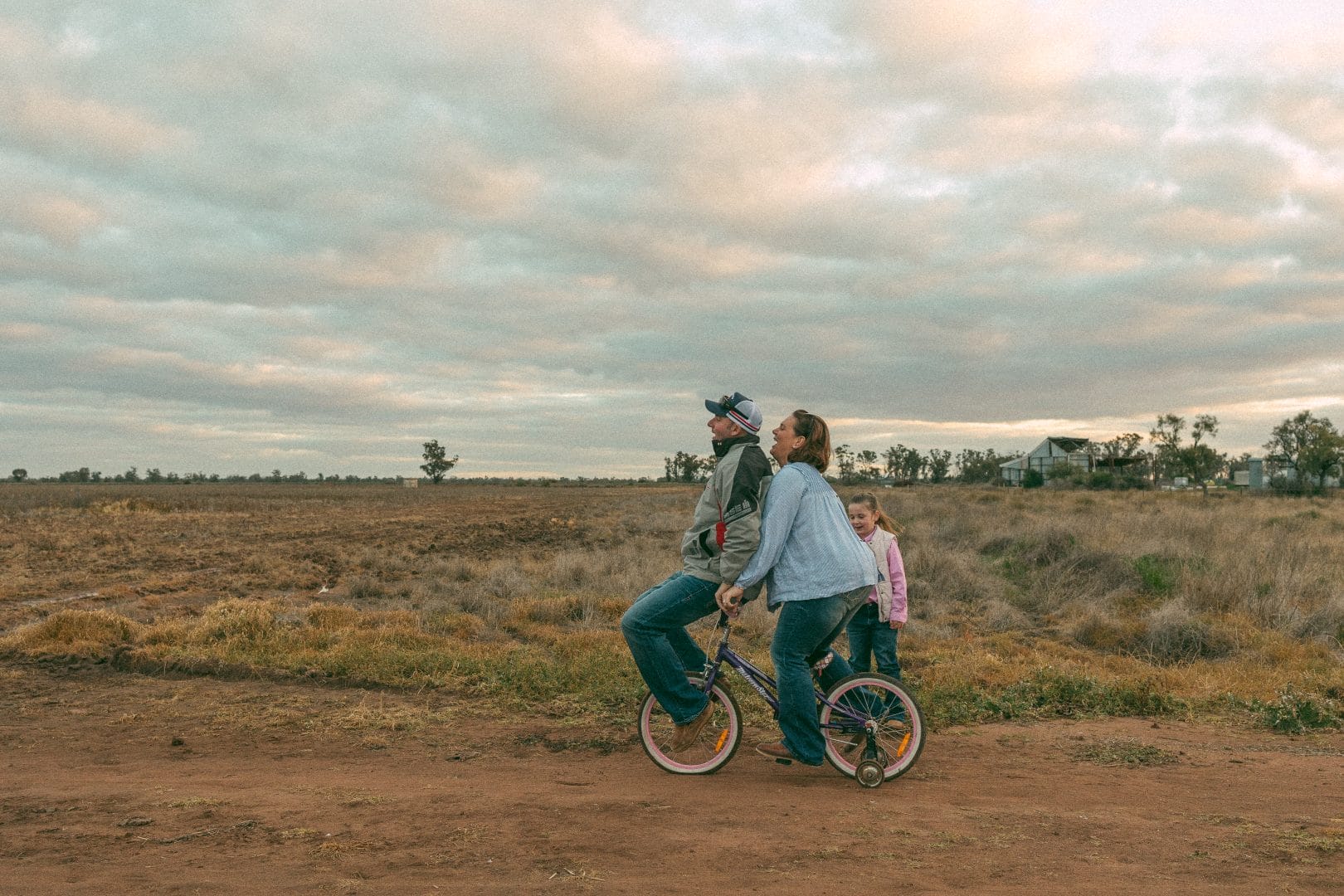 A family of three, with a man and a child on a bicycle and a woman walking beside them, crossing a rural field under a cloudy sky.