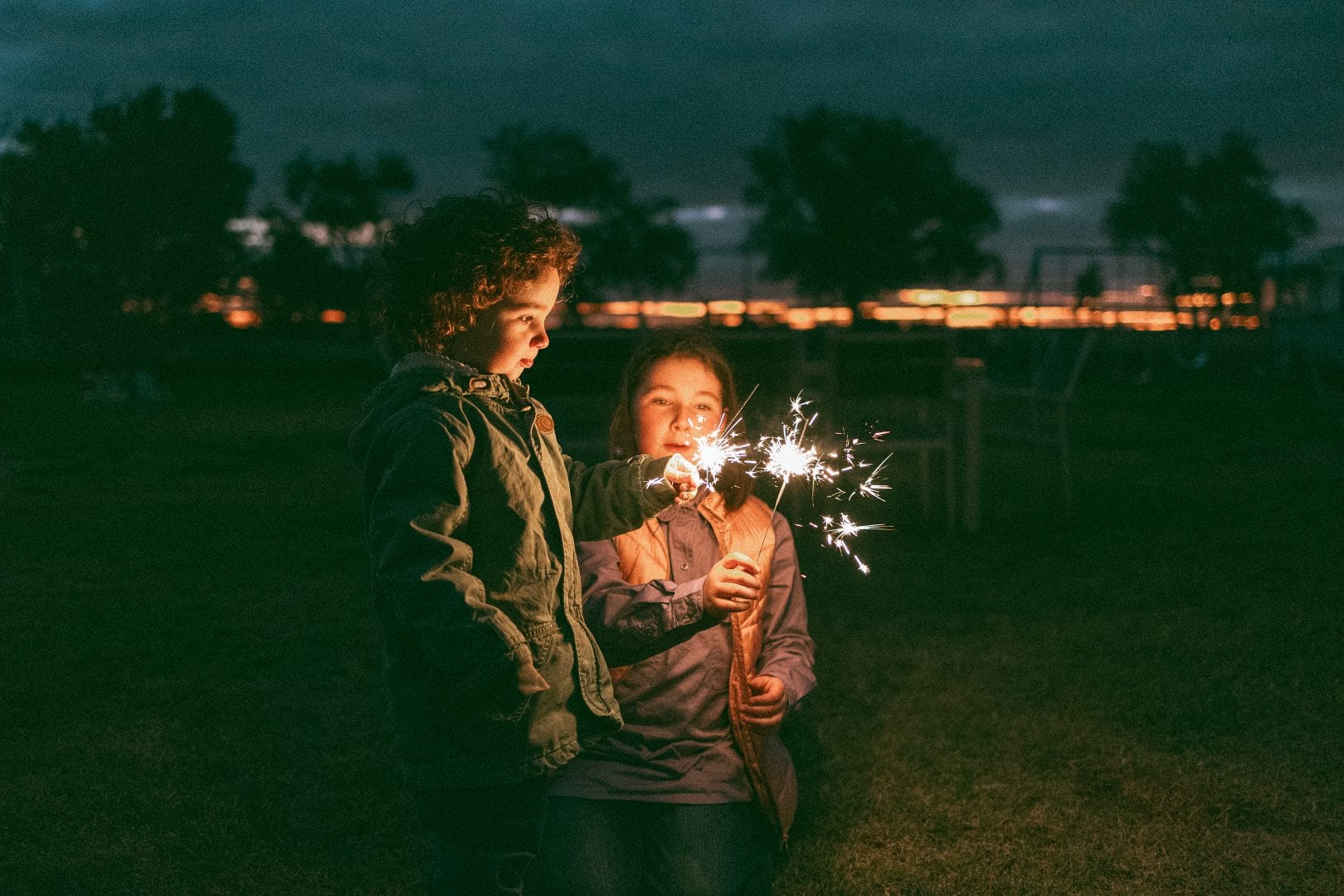 Two children holding sparklers in a field at dusk, with soft focus lights in the background.