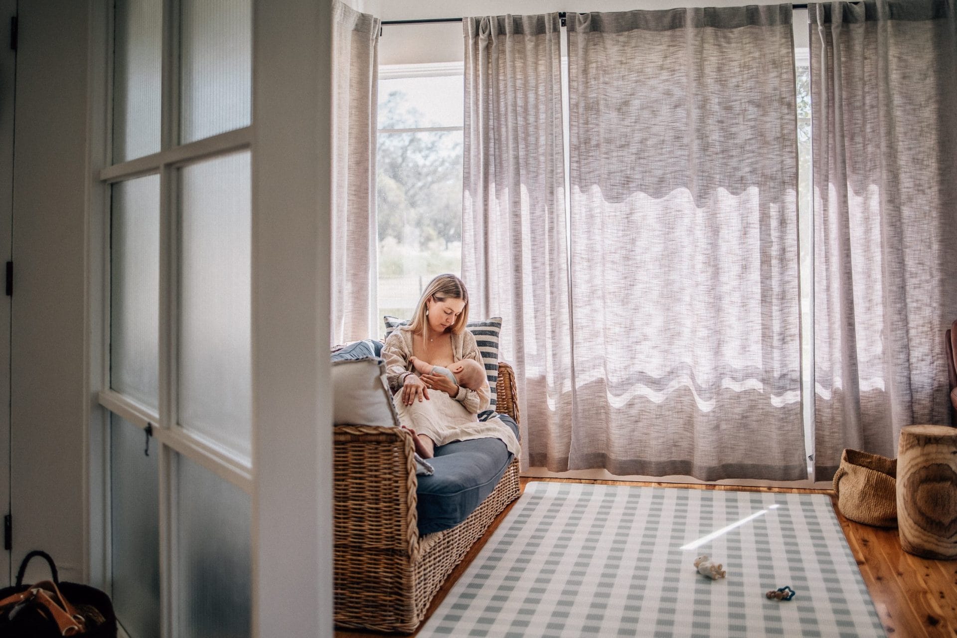A woman sitting in a wicker chair by a window, holding a baby in a cozy, sunlit room with sheer curtains and a checkered rug.