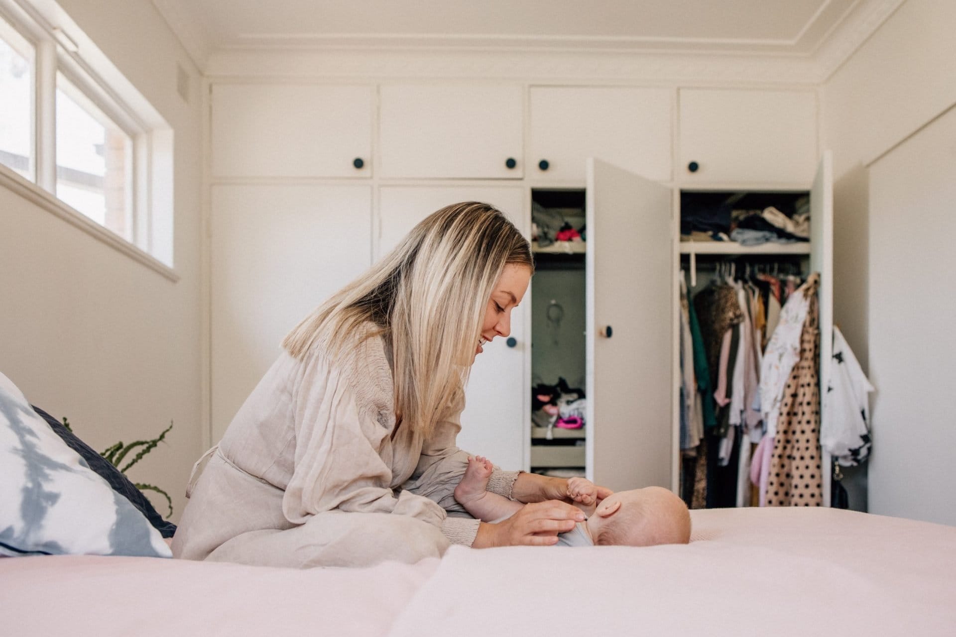 A woman in a cozy sweater gently interacts with a baby lying on a bed in front of open closet doors filled with clothes.