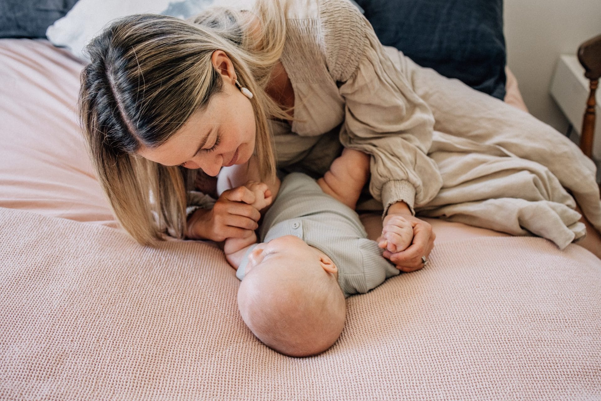 A mother affectionately kisses her baby who is lying on a pink blanket on a bed.