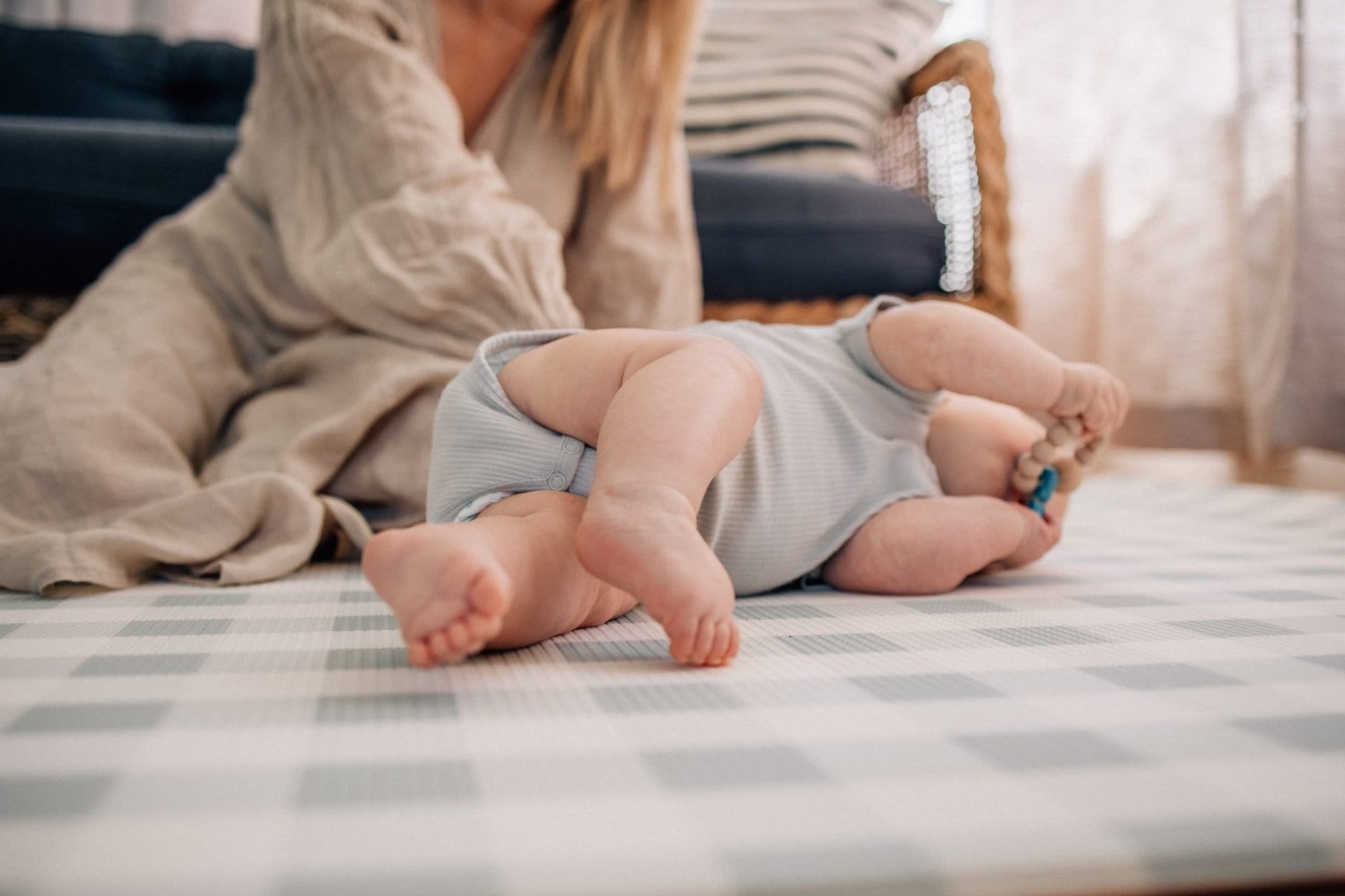 A baby lying on its stomach on a patterned mat, with a woman sitting in the background, partially visible.