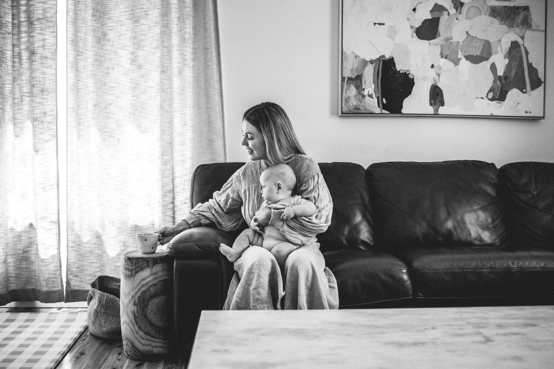 A woman sitting on a couch, holding a baby and looking down, with a cup in her hand, in a cozy living room setting.