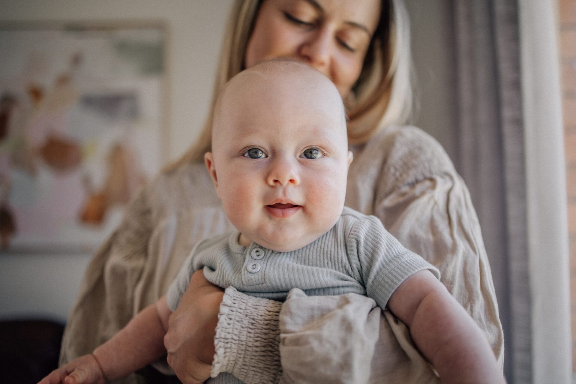 A woman holding a baby with blue eyes; both are partially in focus against a softly blurred background in a cozy room.