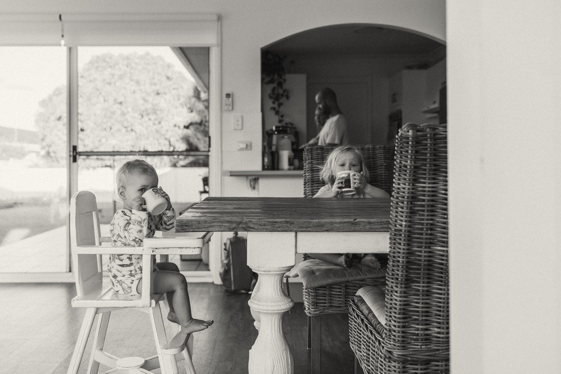 Two toddlers sit at a dining table, one drinking from a cup, with an adult in the background of a home kitchen.