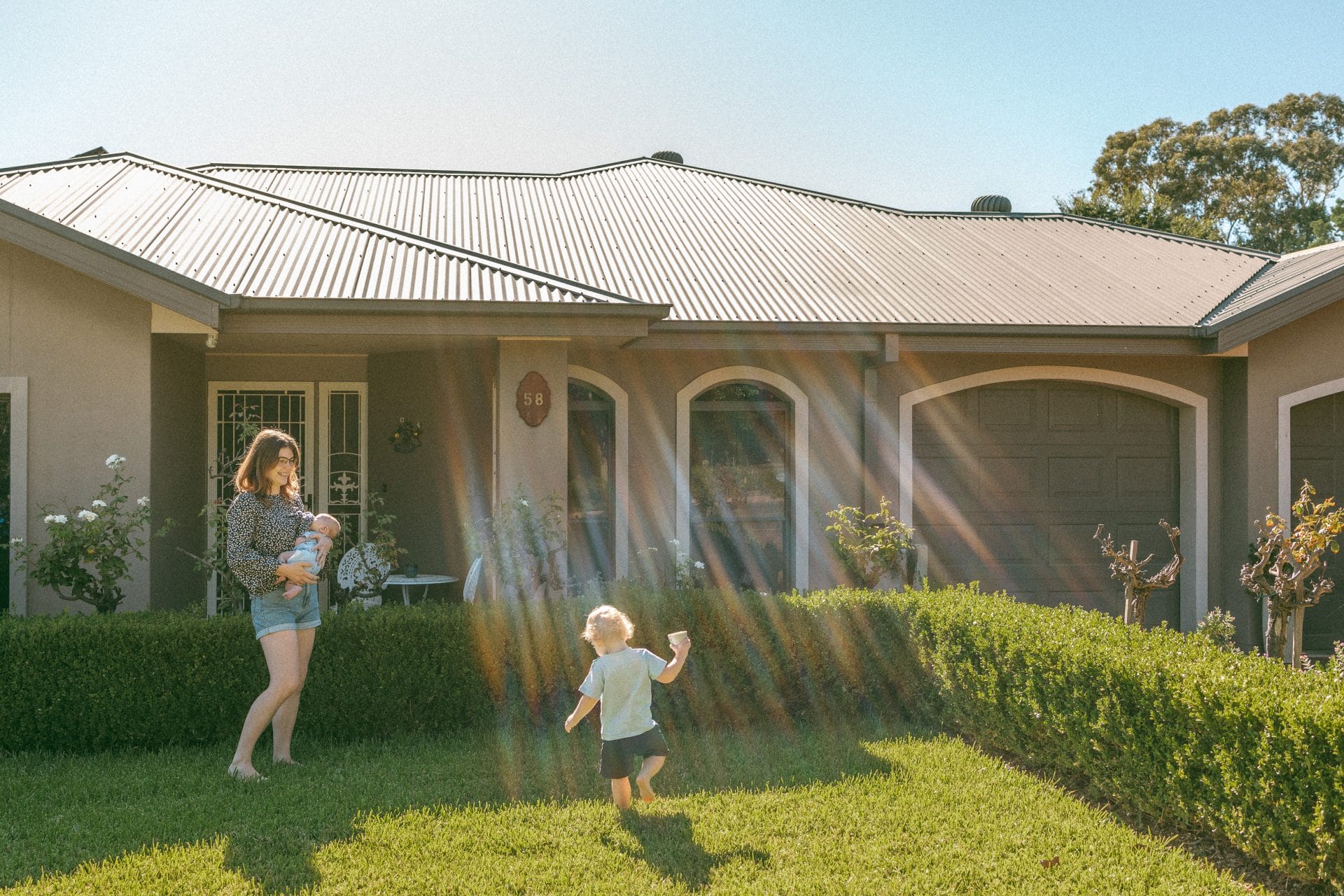A woman and a toddler playing in a sunny yard in front of a house.