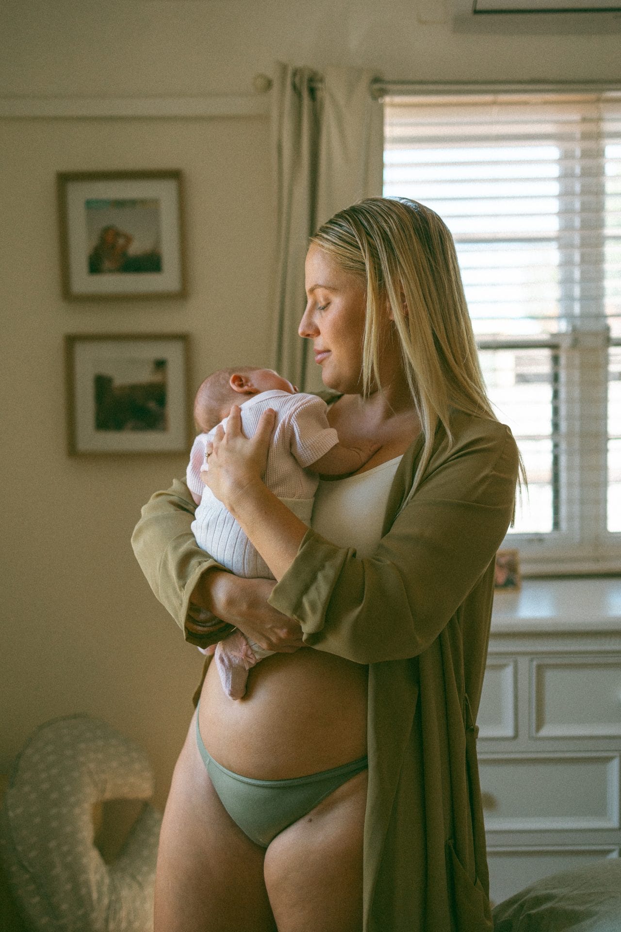 A woman in a beige cardigan and underwear holds a baby in a bright, homey room with framed photos on the wall.