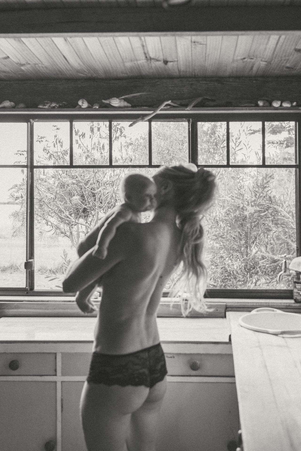 A monochrome image of a woman in undergarments holding a baby, both facing a kitchen window with a view of trees.