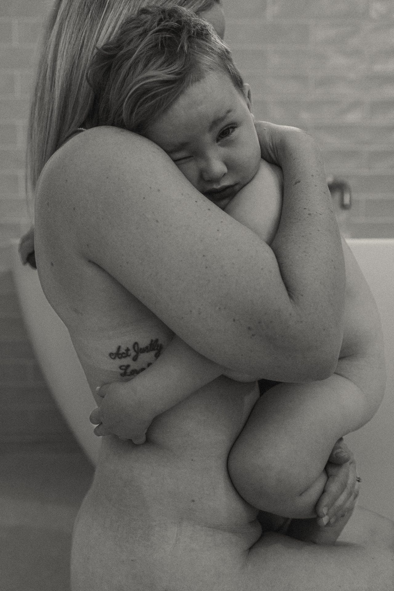 A black and white photograph of a mother embracing her young child, who appears upset. the mother's tattoo "act justly love" is visible on her arm.