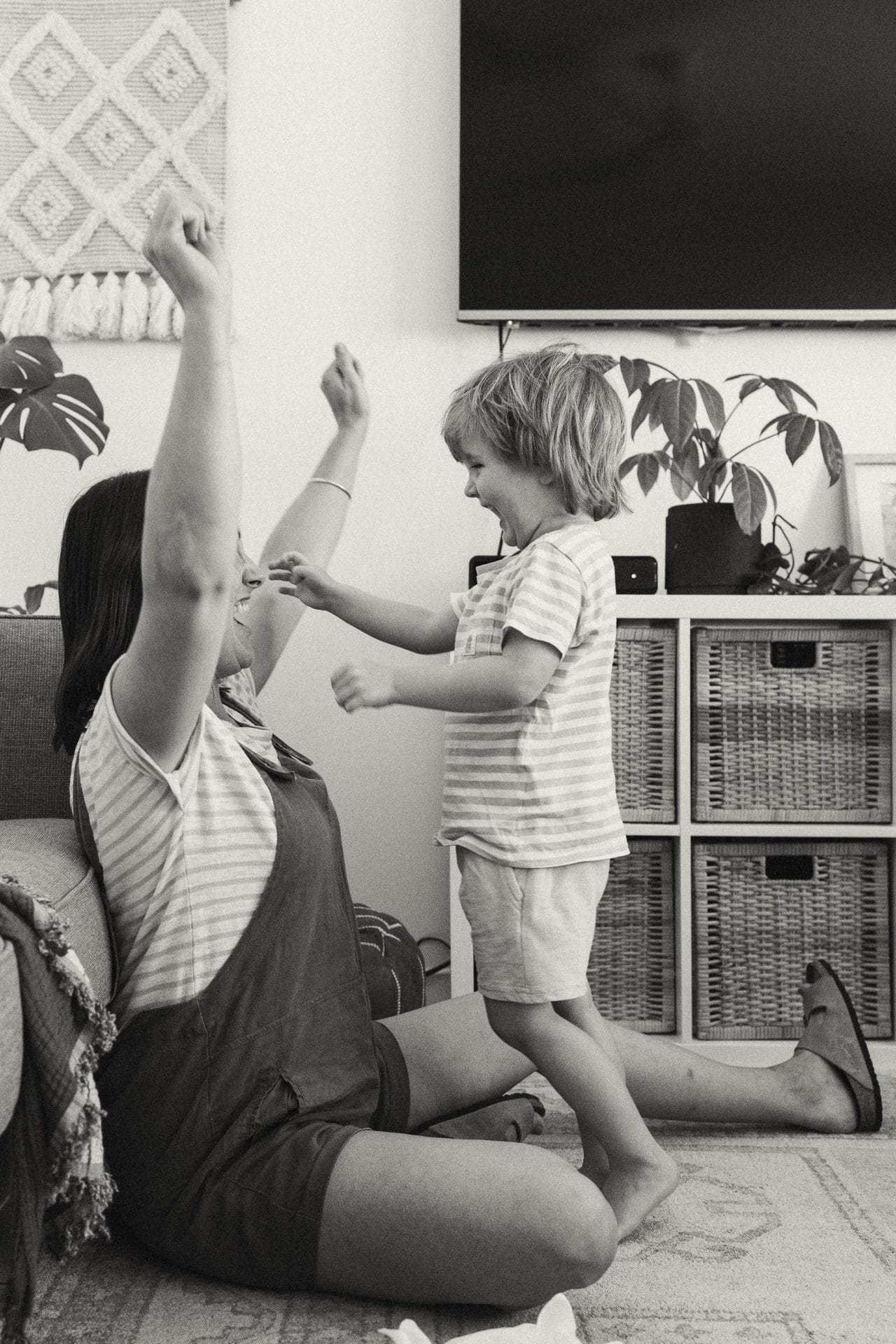 A woman and a young boy playfully raise their hands while sitting on the floor in a living room, surrounded by plants and furniture.