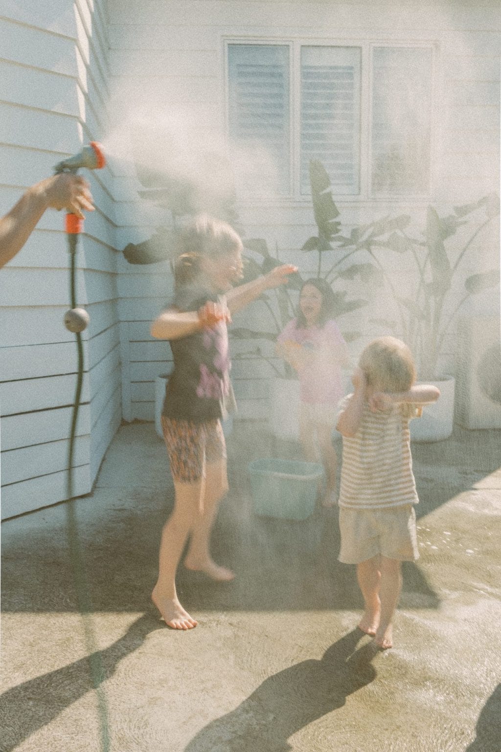 Children playing with water sprayers on a sunny day outside a house, with water droplets illuminated by sunlight.