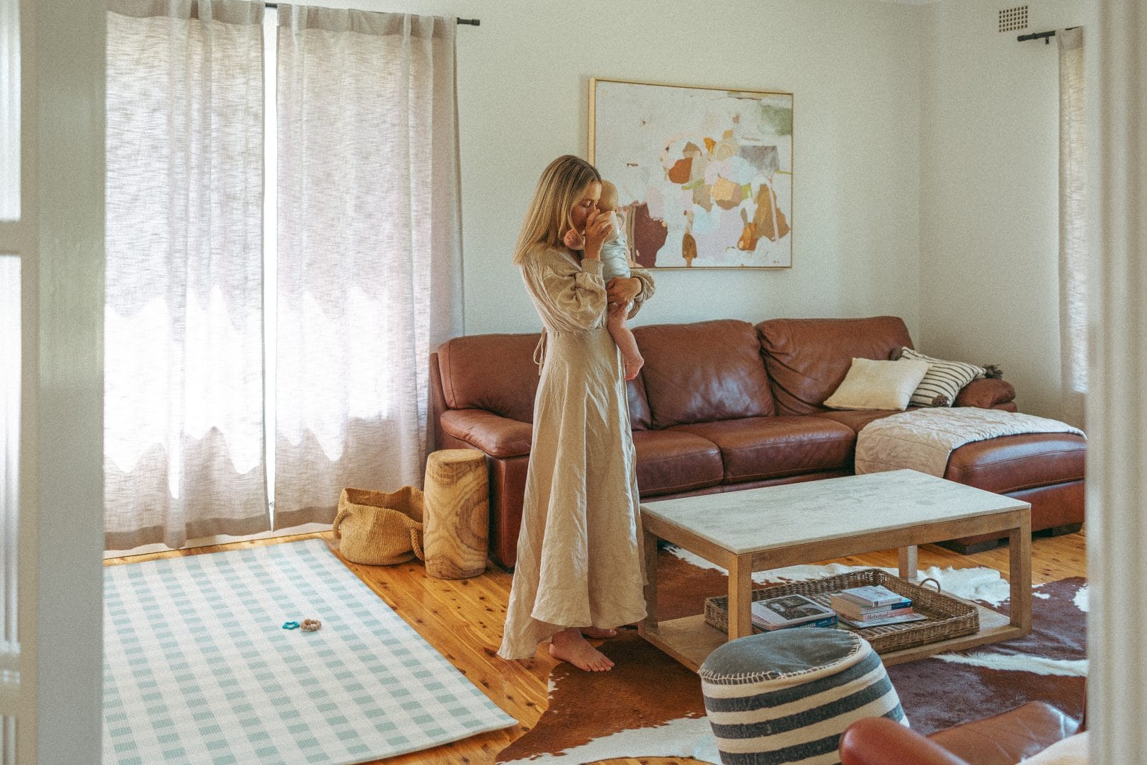 A woman in a long dress holds a baby in a sunlit living room with a brown leather sofa, a white coffee table, and light curtains.