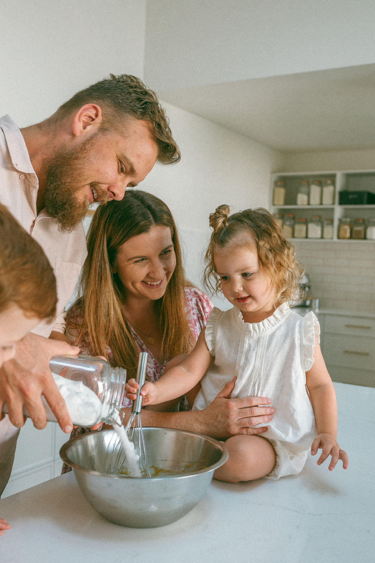 A family of four is gathered around a kitchen counter. The adults assist the children in baking, pouring ingredients into a large mixing bowl. Shelves with jars are visible in the background.