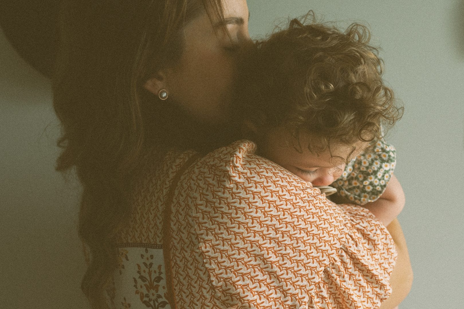 A woman gently kissing the head of her curly-haired toddler, both in soft-focus against a neutral background.