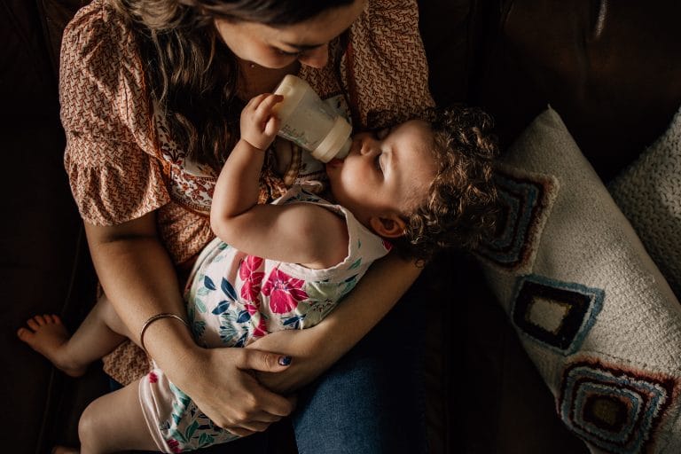 A mother sitting on a couch feeding her toddler from a bottle, both looking at each other affectionately.