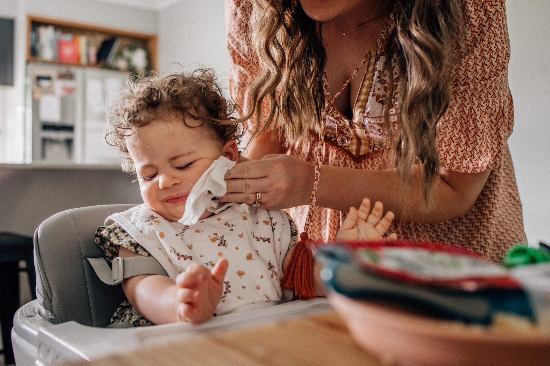 Mother's Day. One day of the year isn't enough. 4 A woman wipes a toddler's face as they sit in a highchair at a dining table, with books and a colorful plate nearby.