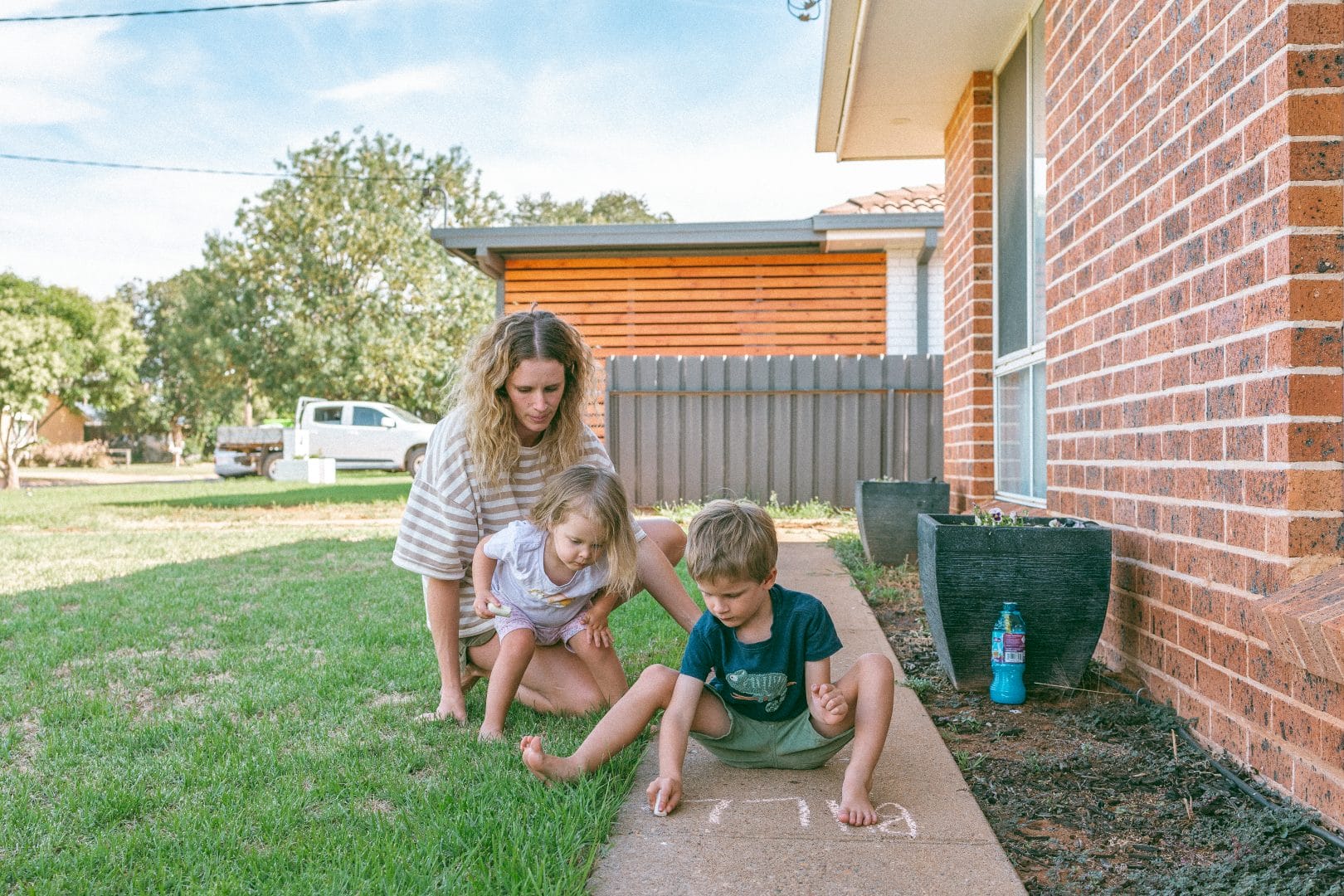 A woman and two children draw with chalk on a driveway next to a brick house, with grass and a tree visible in the background.