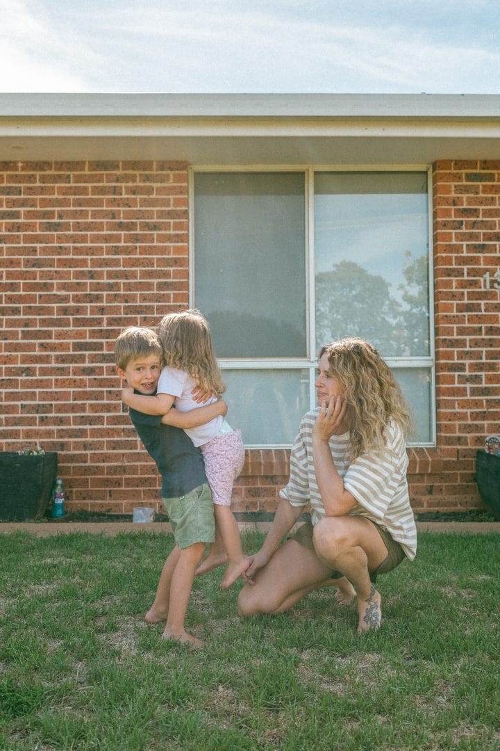 A woman kneeling on grass, smiling at two children embracing each other in front of a brick house with a large window.