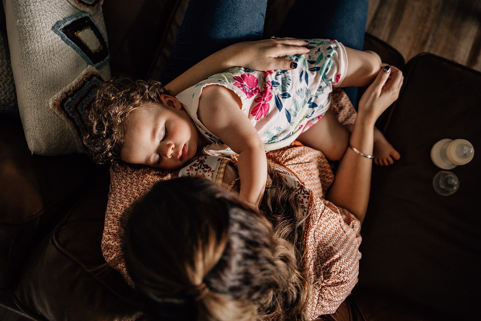 Mother's Day. One day of the year isn't enough. 7 A young child sleeps on a woman's lap on a brown sofa, with a baby bottle nearby. both are indoors and the child wears a floral outfit.