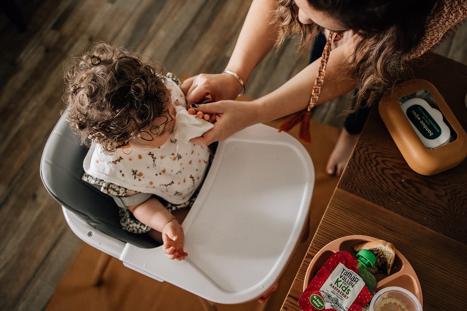Mother's Day. One day of the year isn't enough. 3 A woman adjusts a curly-haired toddler's bib in a high chair, with a tray of food items next to them.