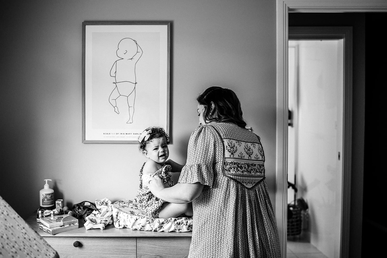 Mother's Day. One day of the year isn't enough. 6 A woman and a young girl smiling at each other, standing in a bedroom with a framed child's drawing on the wall.