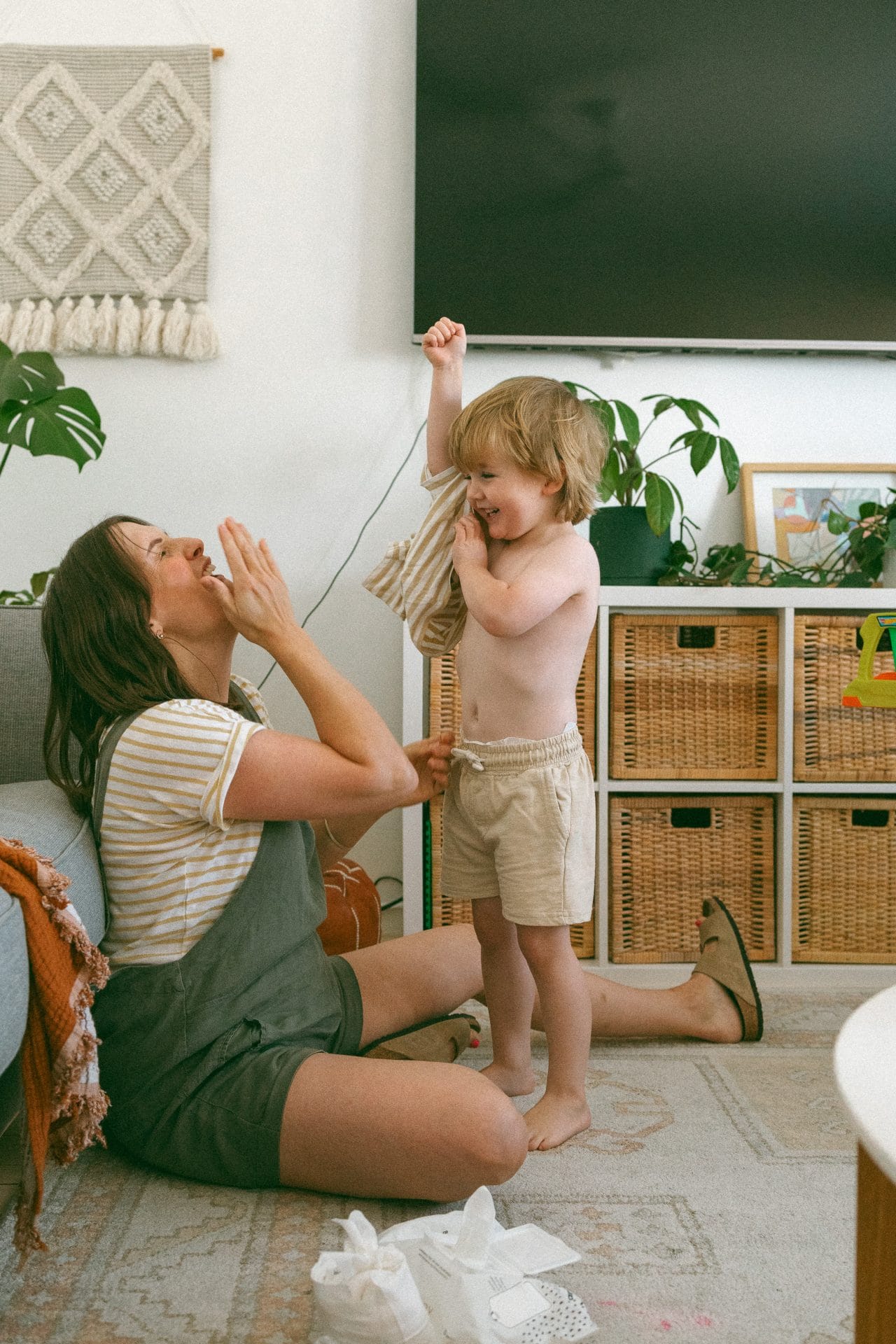A woman plays with a toddler in a living room, using a string to interact while both are smiling joyfully.
